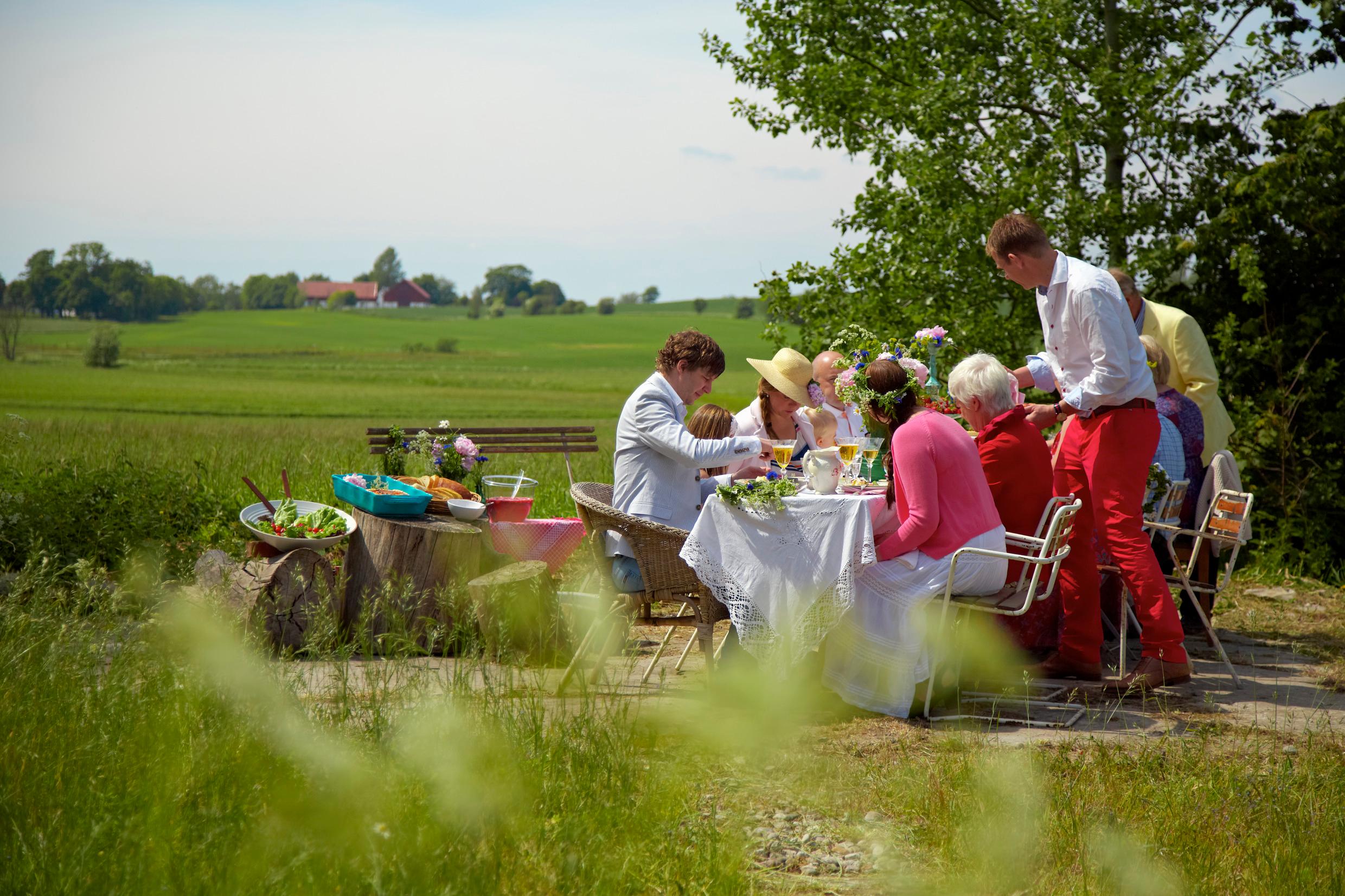 A group of people are sitting down by a table out in a field, eating and drinking. Some wear Midsummer flowers in their hair.