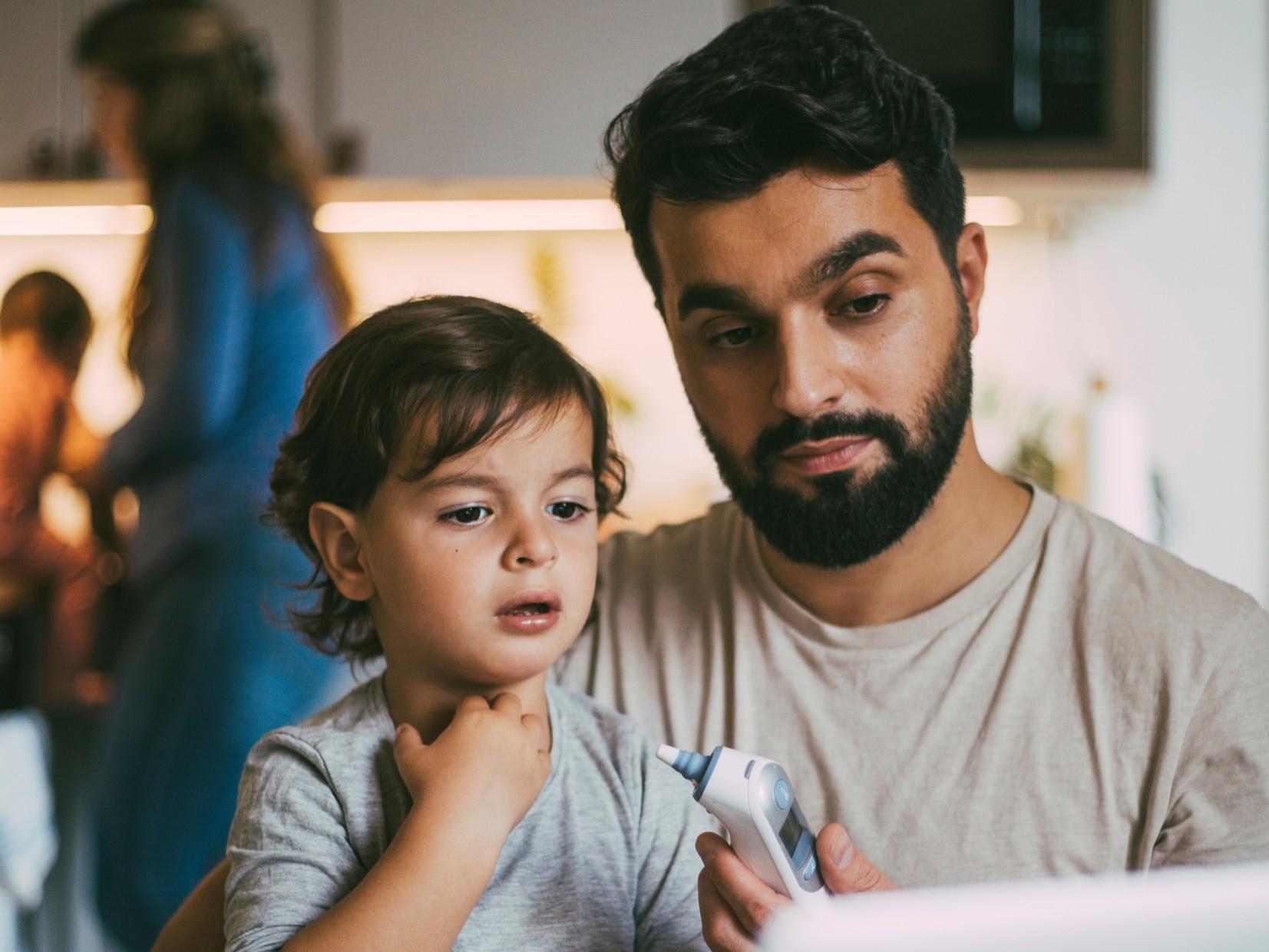 An adult holding a child while checking a thermometer at home.