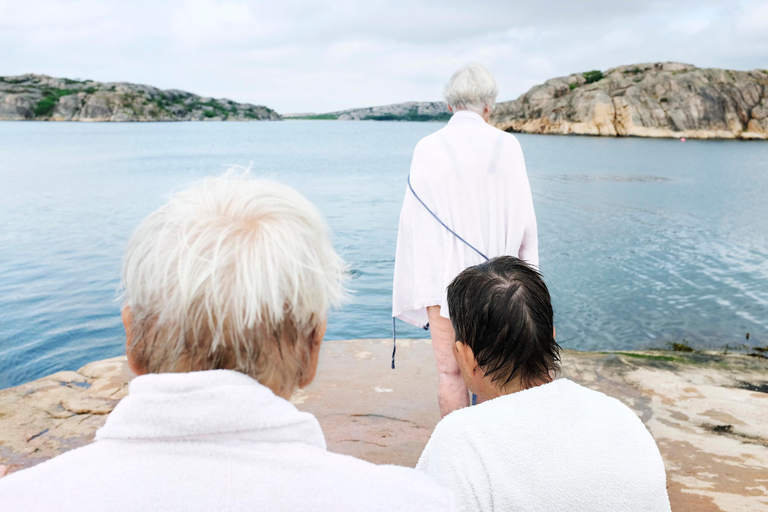 Going for a swim Three elderly women by the sea, all seen from the back and wearing bath robes.