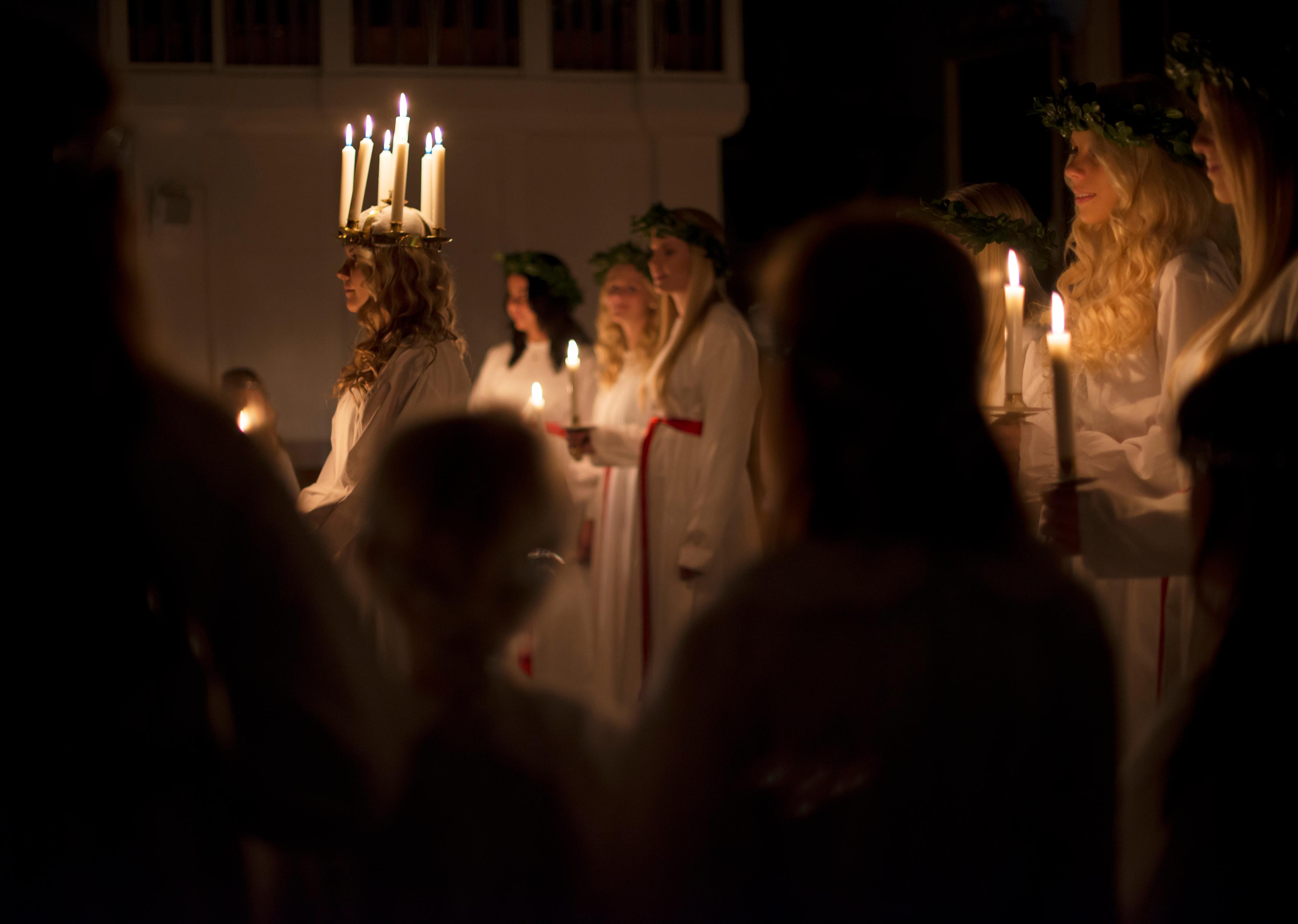 Lucia Day Lucia, with a crown of lit candles on her head, stands in front of the handmaidens who are each holding a lit candle in their hands.
